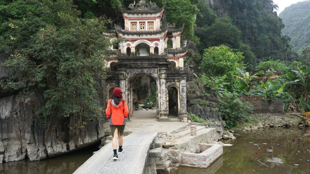 20 Tips from a Local for Planning a Trip to Vietnam. A woman is walking towards an old gate on a narrow bridge over water with lush green mountains in the background in Vietnam.