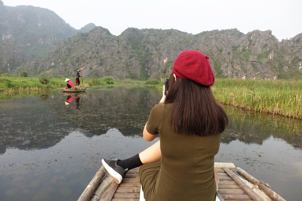 20 Tips from a Local for Planning a Trip to Vietnam. A woman in a red beret is sitting on a bamboo platform taking a photo of two people on a boat in the water with mountains in the background. 