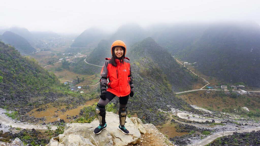 20 Tips from a Local for Planning a Trip to Vietnam. A woman in an orange motorbike helmet wearing a red jacket and protective arm and leg gear is standing on a rock on a cliffside with green mountains in the background. 