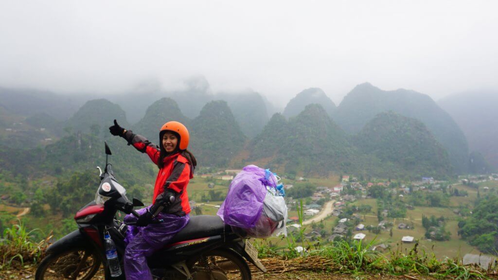 20 Tips from a Local for Planning a Trip to Vietnam. A woman in an orange motorbike helmet, a red jacket, and purple pants is siting on a motorbike with her thumb up and mountains in the background. 