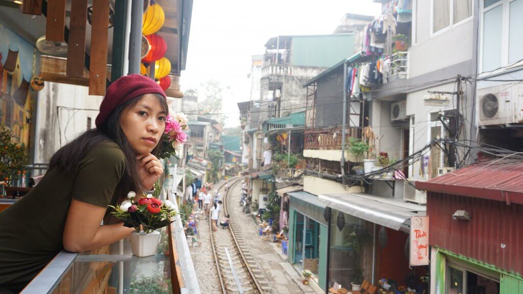 20 Tips from a Local for Planning a Trip to Vietnam. A woman wearing a red beret leans over the edge of a balcony facing the camera. She is overlooking train tracks with homes on either side of the tracks and people walking along the sides and on the train tracks. 