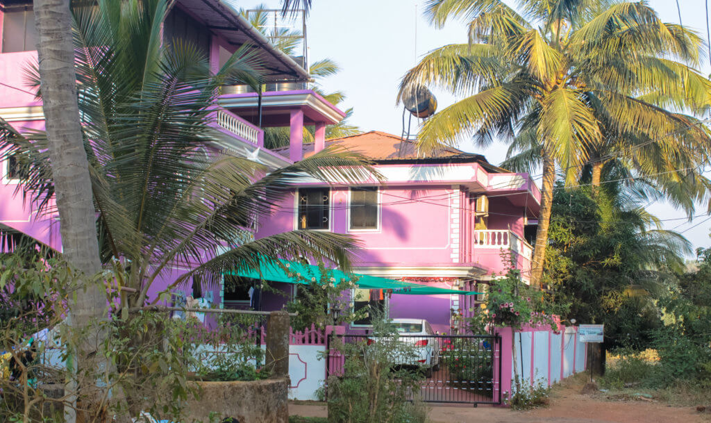 long-term guesthouse stay in India. Picture is of a purple two story house with palm trees on either side. 