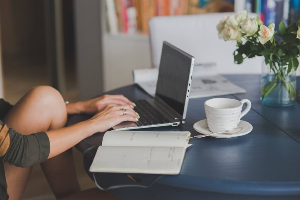 How to Become a Digital Nomad. Your Ultimate Guide to starting Digital Nomad life. A person is sitting with their knees against a blue table working on an open laptop. They have an open notebook and cup of coffee next to them. There are white flowers on the table. 