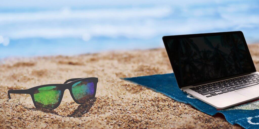 How to Become a Digital Nomad. Your Ultimate Guide to starting Digital Nomad life. A laptop is open on top of a beach towel on the sand in front of the ocean. There is a pair of sunglasses on the sand next to it. 