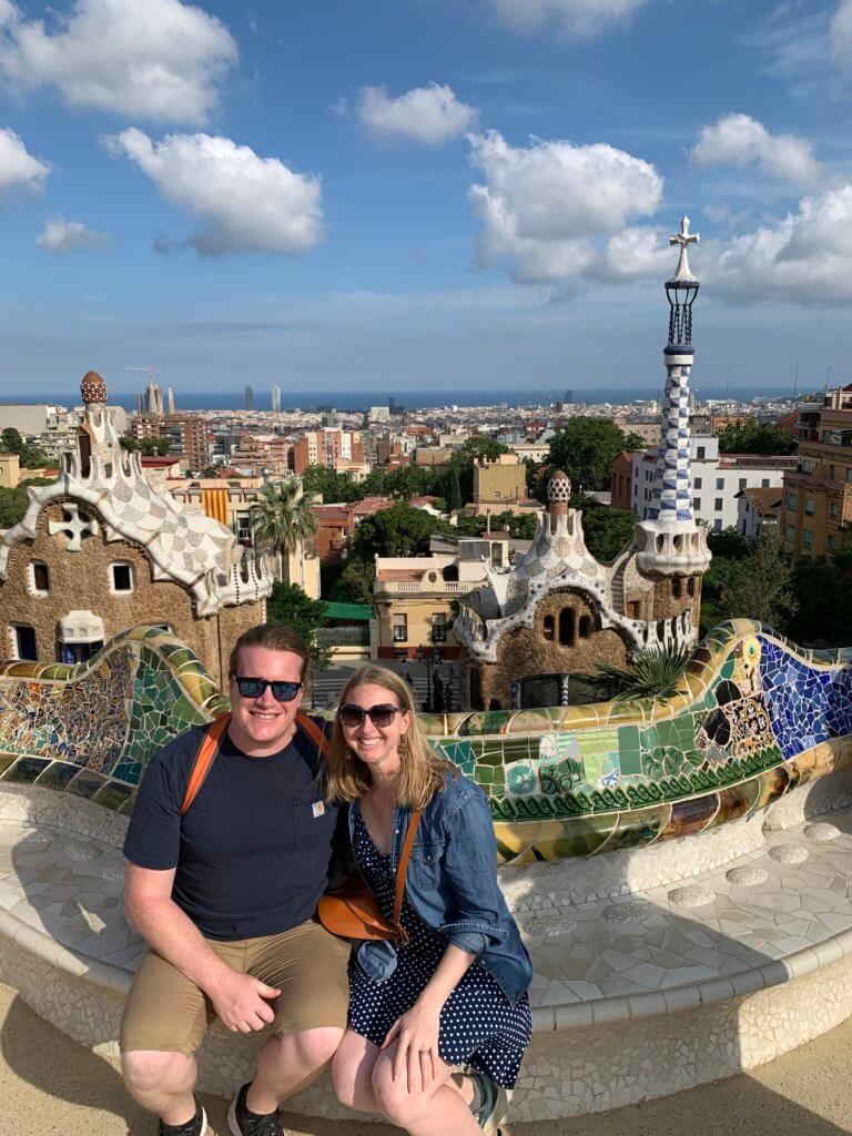 Gaudi Buildings in Barcelona Jen and Stevo sitting on the mosaic tile bencg at Park Guell looking out over the city