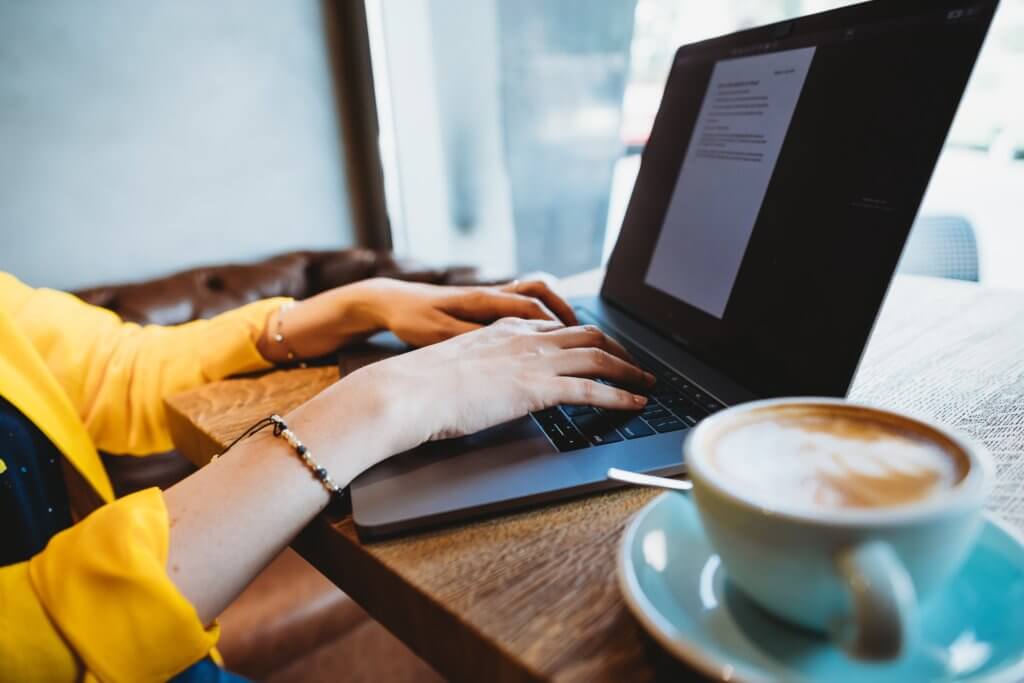  A woman in a yellow shirt is typing on her laptop with a blue cup of coffee next to her. 