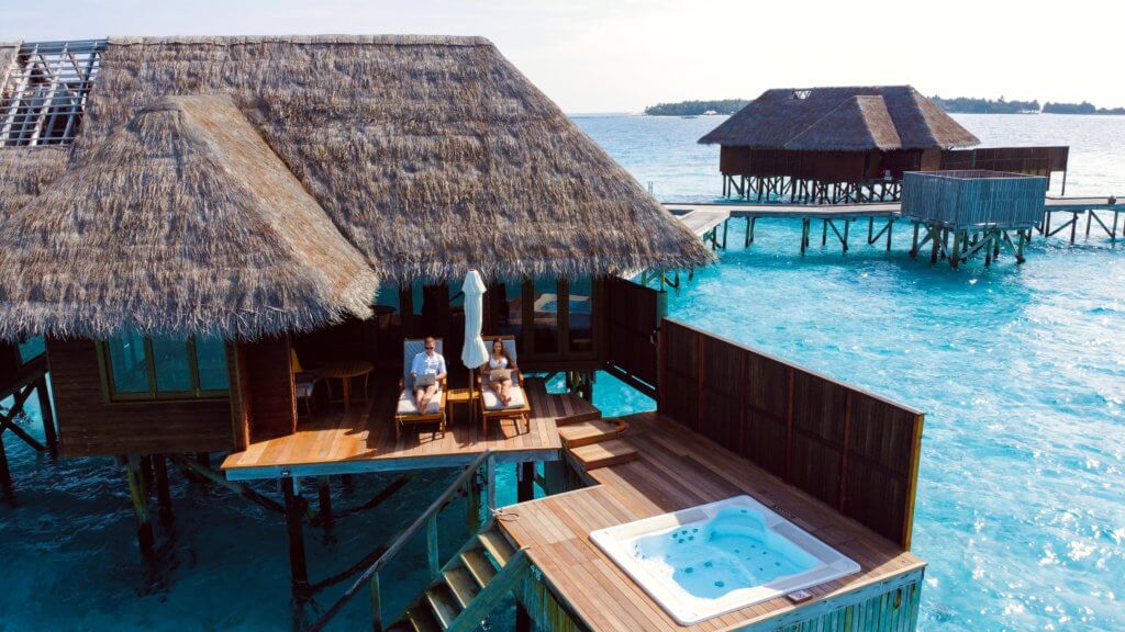 A couple is sitting on the terrace of their overwater bungalow. Shot is taken from a drone. You can get a digital nomad visa to stay long term in places like this!