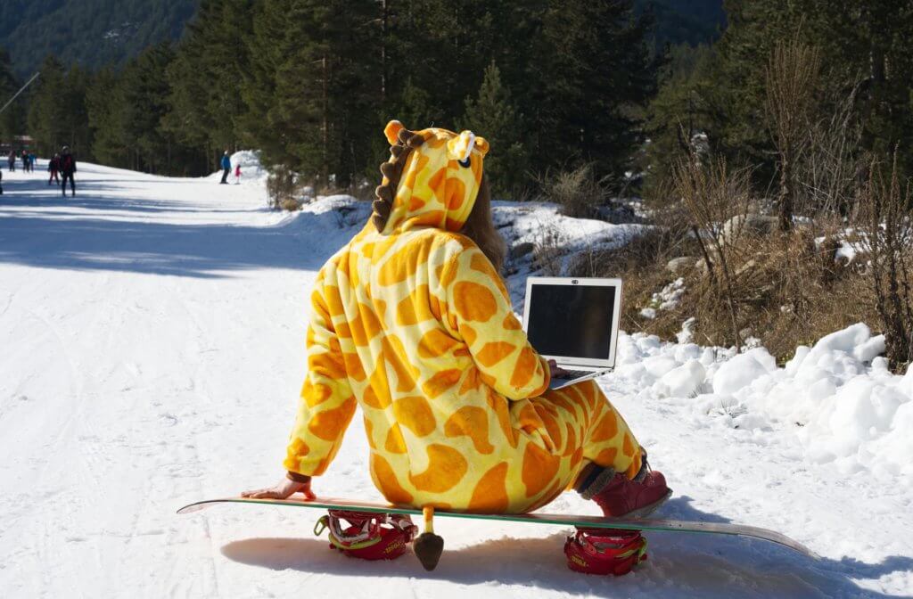 A woman wearing a giraffe onesie is sitting on an upside-down snowboard with her back to the camera and a laptop on her lap in the middle of a ski run with other skiers and snowboarders riding past her. 
