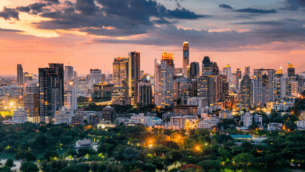 A photo of Bangkok's city skyline at dusk with lights coming on in the tall buildings and a park in the foreground.